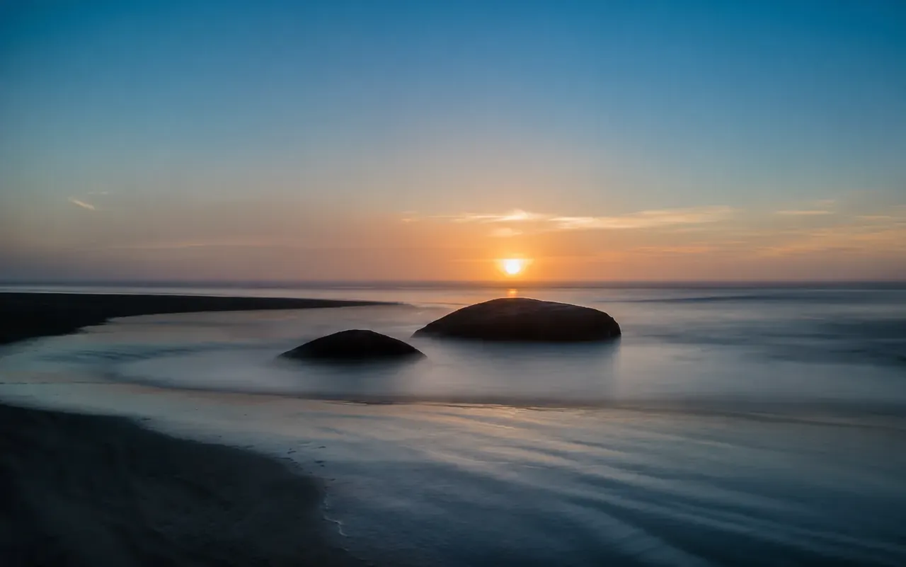 Long-exposure coastal sunset with dark rocks in soft-moving water